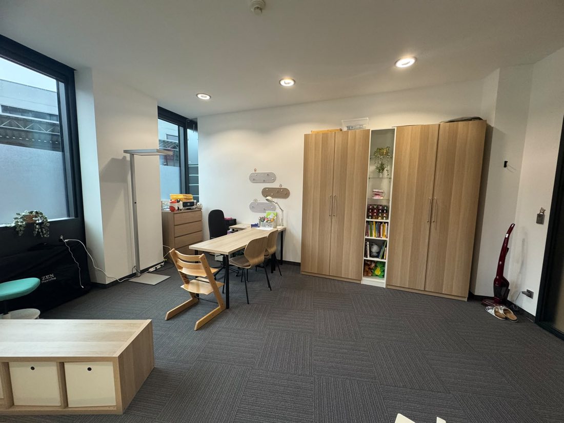 Der Arbeitsbereich desselben Raums: ein heller Holztisch mit Stühlen,
                    darunter ein mitwachsender Kinderstuhl, daneben Schränke und ein
                    offenes Regal mit Büchern und Spielsachen.
